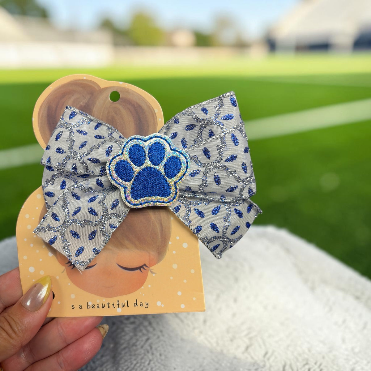 Mloved Designs silver and blue glitter hair bow with an embroidered paw print patch on a display card.