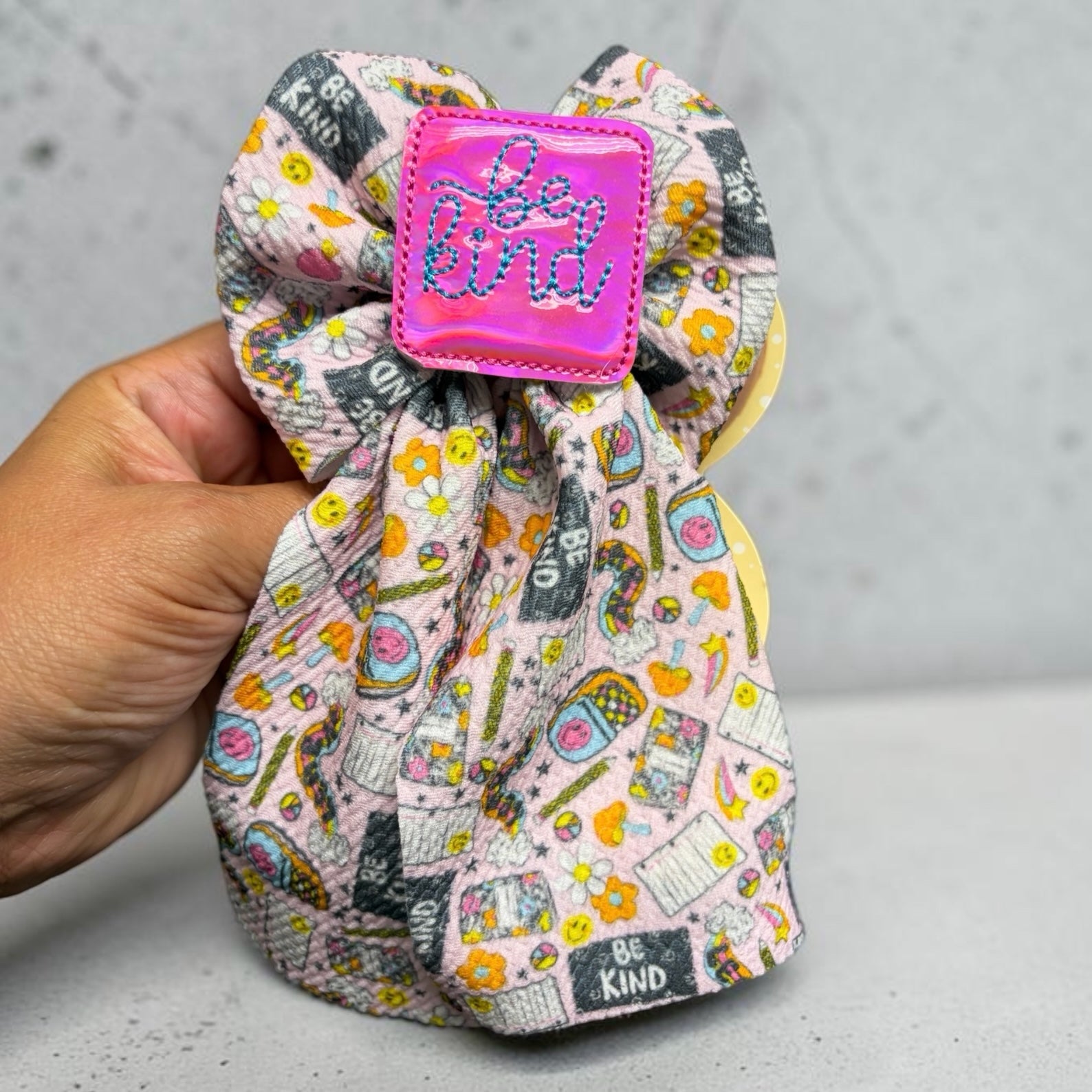 Mloved Designs fabric hair bow with a colorful floral and smiley face print and a pink holographic be kind patch.