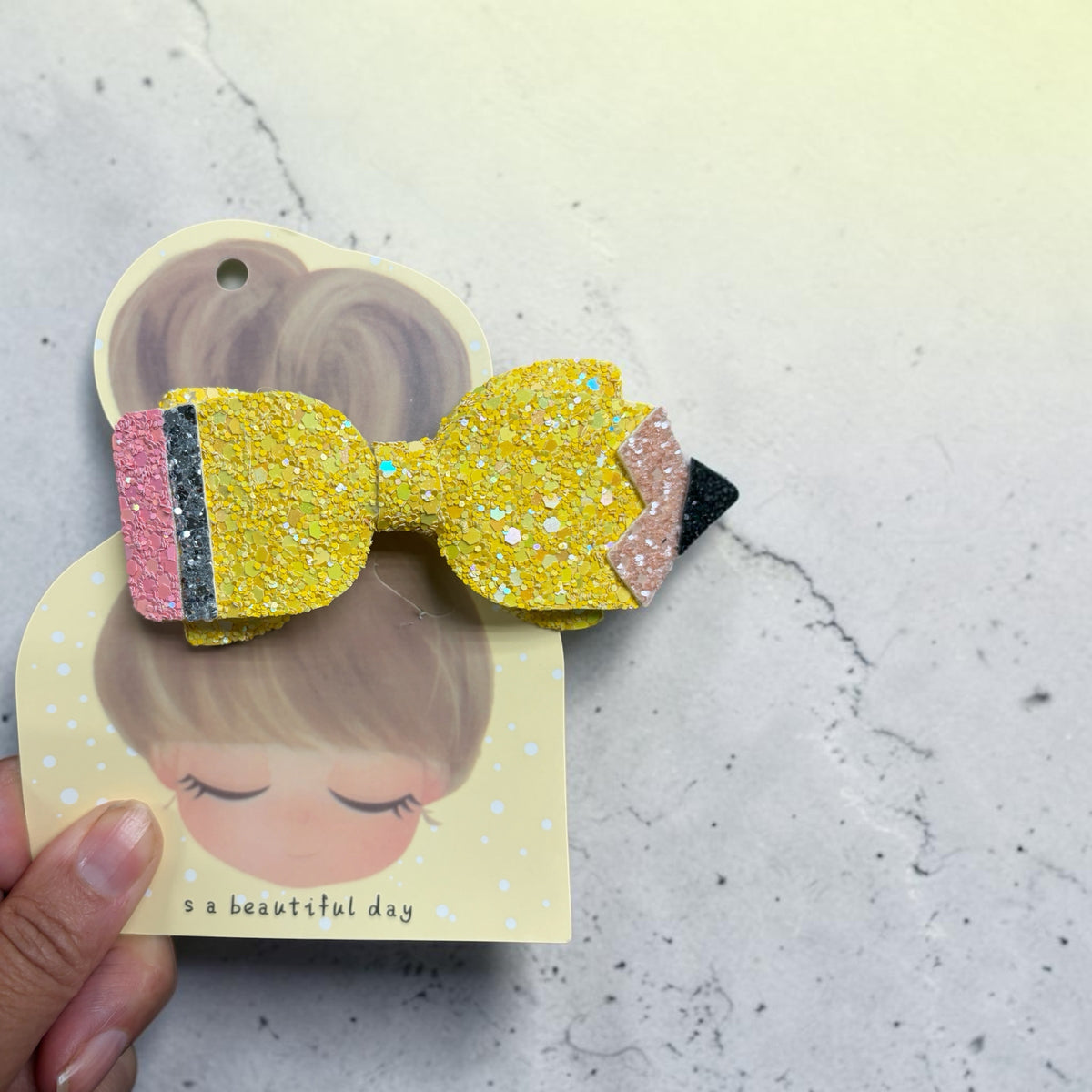Mloved Designs pencil-shaped hair bow made of chunky yellow, pink, and black glitter fabric on a display card.