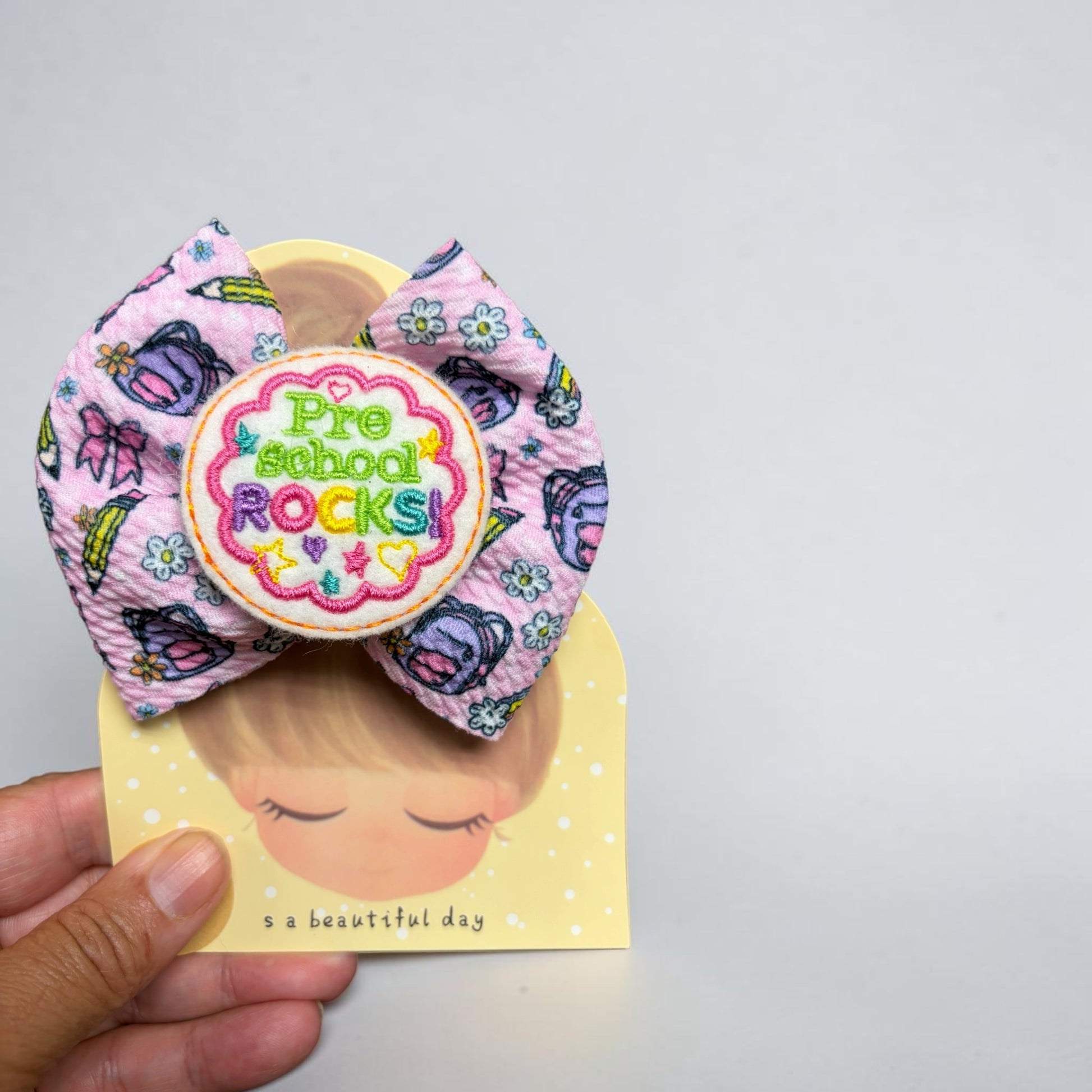 Mloved Designs pink hair bow with school supply pattern and embroidered Pre-School Rocks patch on display card
